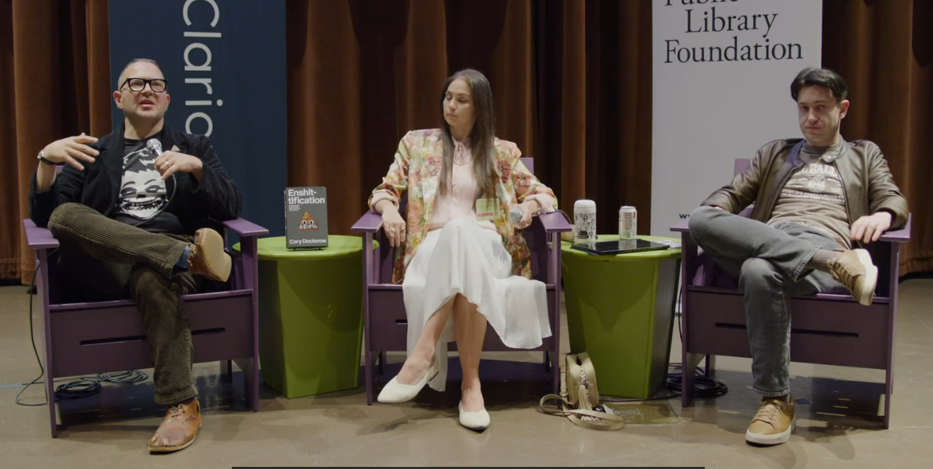 A stage at the Seattle Public Library; on it sit Ed Zitron, Whitney Beltran and Cory Doctorow, holding mics.