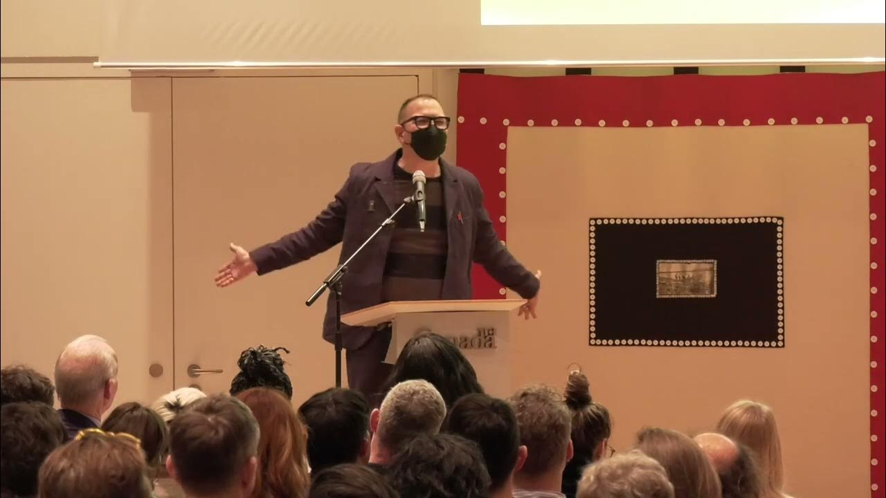 A photo of me, wearing a suit and a mask, standing with my arms open at a podium at the Canadian embassy in Berlin, giving my McLuhan Lecture.
